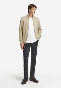 Ung man iklädd en beige zip-up cardigan, vit t-shirt, mörka slim-fit byxor och svarta läderskor, står med ena handen i fickan.