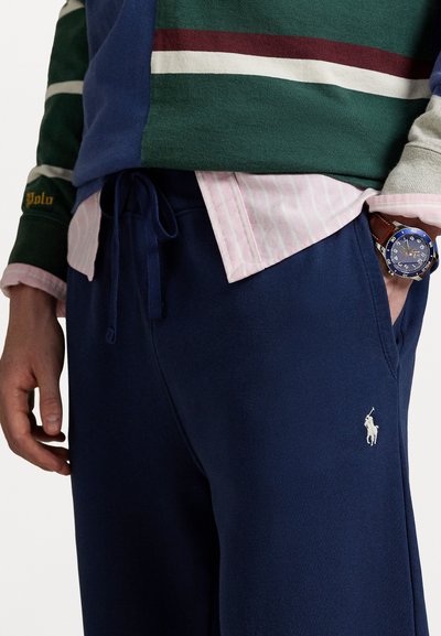 Mann som har på seg marineblå joggebukser med polo-logo, stripete rosa skjorte, grønn og burgunderstripete genser, og blå klokke med brun lærrem.
