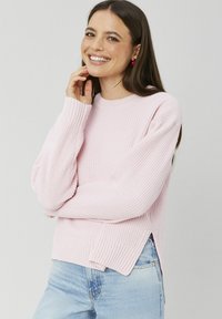 Pull en tricot rose clair avec une texture côtelée, encolure ronde, fentes sur les côtés et manches longues. Assorti à un jean bleu.