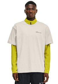 Jasno beżowa krótki rękaw koszulka nałożona na żółtą koszulę z długim rękawem, z czarnym nadrukowanym logo po lewej stronie klatki piersiowej.