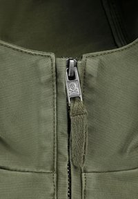 Tissu vert olive avec une surface texturée, comportant une fermeture éclair en métal argenté avec un emblème de crâne et os croisés sur le tirette.