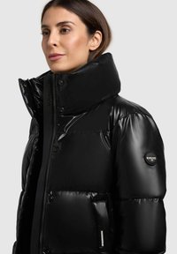 Juodas pūkinis švarkas su aukštu apykakle, blizgiu paviršiumi, sagomis ir logotipo žyme kairiajame rankovės pusėje. Su šoninėmis kišenėmis.