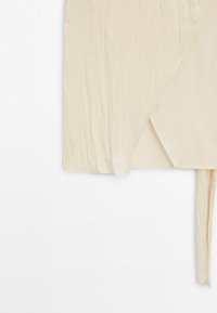 Haut cache-cœur à manches longues de couleur crème avec tissu texturé et détail noué à la taille sur fond blanc.