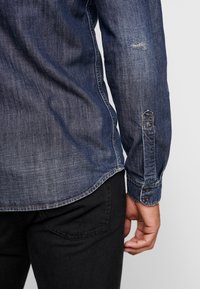 Camicia in denim con una finitura blu scuro, caratterizzata da sbiaditure visibili, chiusura a bottone sui polsini e orlo curvo. Indossata con jeans neri.
