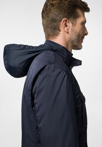 Granatowa wodoodporna kurtka z wysokim kołnierzem i kapturem. Posiada napy oraz boczną kieszeń na piersi. Gładka, matowa tekstura.