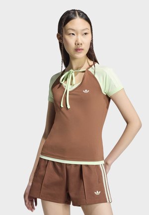 Jonge vrouw met gevlochten haar draagt een bruine en lichtgroene Adidas sporttop en bijpassende bruine korte broek met witte strepen, staand met hand op de heup.