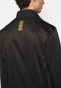 Giacca nera realizzata in materiale liscio, caratterizzata da una stampa a strisce dorate sul retro e da un colletto classico. Leggera con una silhouette elegante.