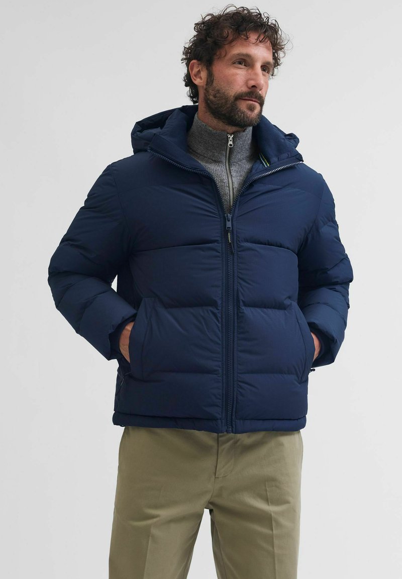 Giacca puffer blu navy con cappuccio, dotata di zip frontale e tasche angolate. Il materiale sembra liscio, con sezioni trapuntate e squadrate.