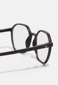 Lunettes noires à monture octogonale avec des verres transparents, dotées d'une finition mate et d'un petit logo sur la branche.