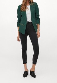 Grön skräddarsydd blazer med en elegant design, två framsidans dragkedjor, parat med svarta slim fit-jeans och svarta öppna clogs.