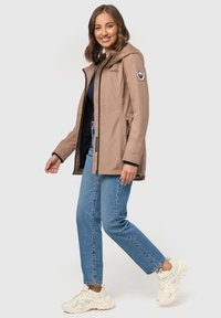 Beige waterproof jacket with a hood, side zippers, and logo on the sleeve, paired with blue jeans and white sneakers.