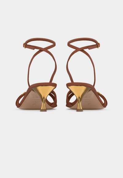 Καφέ σανδάλια με λεπτά λουριά στον αστράγαλο και λαμπερά χρυσά τακούνια γατούλας, απεικονιζόμενα από πίσω σε απλό φωτεινό φόντο.