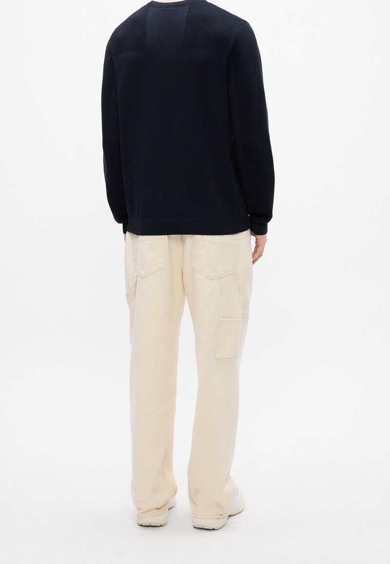 Personne portant un t-shirt noir à manches longues et un pantalon cargo beige ample, debout de dos sur un fond blanc.