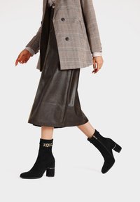 Stivali neri alla caviglia con tacco a blocco, realizzati in suede, con un accento dorato e una cerniera, abbinati a una gonna in pelle marrone e a un blazer a quadri.