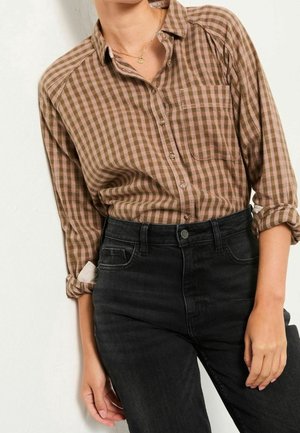 Personne portant une chemise à carreaux marron avec les manches retroussées, rentrée dans un jean noir taille haute, debout contre un mur clair.