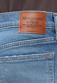 Džínsy z denimu v svetlomodrej farbe s hnedou koženou nášivkou s vyrazeným textom a bežným prešívaním pozdĺž pásu a vrecka.