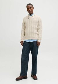 Beige Strickpullover mit Schalkragen, kombiniert mit einem hellblauen Hemd darunter und dunklen, weit geschnittenen Jeans. Braune Lederschuhe.
