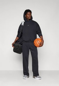 Camisola de capuz preta com bolso frontal, calças de fato de treino combinando e ténis cinzentos. O modelo segura uma bola de basquetebol laranja e uma mala preta com alça.