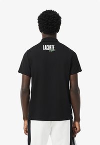 Polo noir avec un col classique. Présente un logo "LACOSTE" blanc et un graphique de crocodile vert au dos. Fabriqué en tissu doux et respirant.