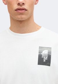 Κοντινό πλάνο ενός ατόμου που φοράει λευκό T-shirt με ασπρόμαυρο λογότυπο The North Face στο αριστερό πάνω μέρος του στήθους.