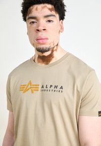 Šviesiai ruda medvilninė t-marškiniai su juodu ir auksiniu "ALPHA INDUSTRIES" logotipu, apvaliu kaklu, trumpomis rankovėmis ir mažuoju logotipu ant rankovės.