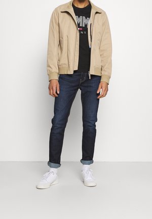 Bombers beige avec fermeture éclair à l'avant, t-shirt graphique noir, jeans bleu foncé et baskets blanches. Tissu lisse et coupe décontractée.