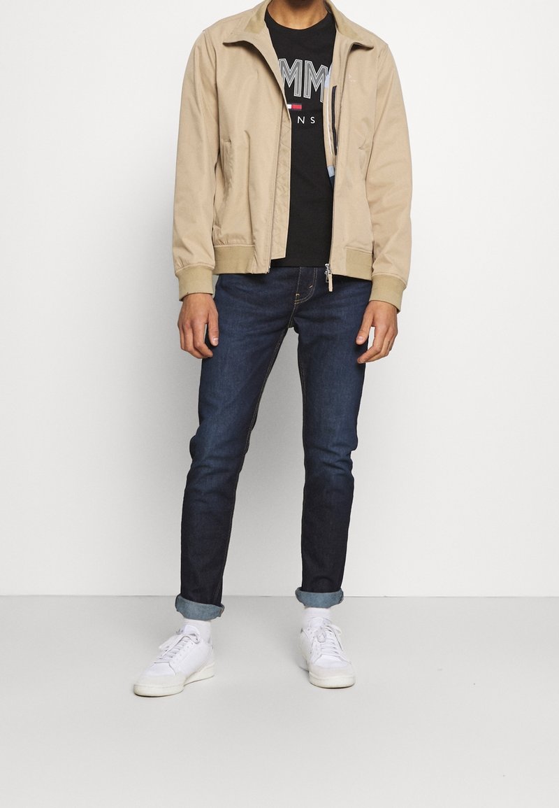 Beige Bomberjacke mit Reißverschluss vorne, schwarzes Grafik-T-Shirt, dunkelblaue Jeans und weiße Sneaker. Glatter Stoff und lässige Passform.