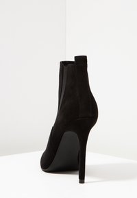 Černé semišové kotníkové boty s vysokým stiletto podpatkem, elastickými bočními panely a hladkým, zaobleným designem špičky. Minimalistický styl s elegantní siluetou.