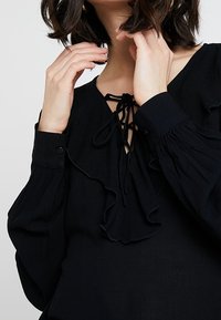 Blusa nera con accenti arricciati, scollatura con coulisse e maniche lunghe plissettate. Tessuto morbido e fluido con dettaglio a bottone sui polsini.