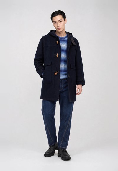 Mann trägt einen marineblauen Dufflecoat über einem blau gestreiften Pullover, blaue Jeans und schwarze Stiefel, steht vor einem schlichten hellen Hintergrund.