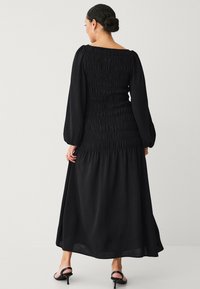 Schwarzes langärmliges Kleid mit gerafftem Oberteil, quadratischem Ausschnitt und fließendem Rock. Aus glattem Stoff mit betonter Taille.