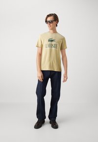 Una camiseta de algodón color caqui presenta un gráfico de "LACOSTE" y el logo del cocodrilo; se combina con jeans oscuros y zapatos negros con cordones.