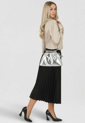 Sac à main en métal argenté avec un dessus froncé et une bandoulière chaîne, associé à une jupe plissée noire et un chemisier beige. Chaussures noires à talons hauts.