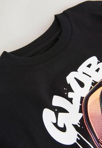 Schwarzer Pullover mit geripptem Rundhalsausschnitt, mit einem großen weißen und orangefarbenen Grafikdesign auf der Vorderseite. Weiche Stofftextur.