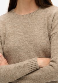 Beżowy wełniany sweter z okrągłym dekoltem i ściągaczami na rękawach. Miękka faktura i luźny krój widoczne na skrzyżowanych rękach.