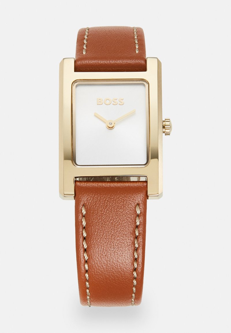 Montre rectangulaire dorée avec un cadran blanc, un bracelet en cuir marron et des coutures claires. Présente des aiguilles dorées minimalistes et une couronne.