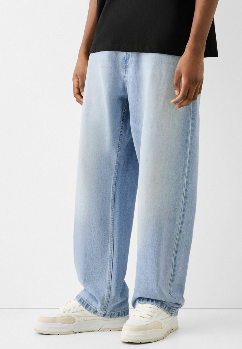 Light blue denim jeans with a relaxed fit, featuring a straight leg design and subtle fading. Paired with white sneakers and a black shirt.