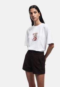 Witte oversized t-shirt met een teddybeer graphic, gecombineerd met hooggerande donkerbruine shorts. Gladde katoentextuur en een relaxte pasvorm.