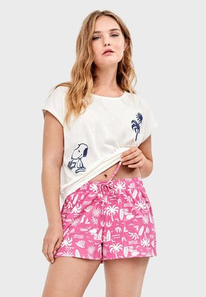 Junge Frau hebt ein weißes T-Shirt mit Snoopy-Aufdruck hoch, trägt pinkfarbene Shorts mit weißen tropischen Mustern und Snoopy-Motiven.