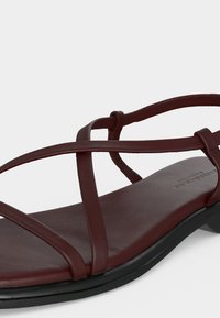 Sandale plate en cuir marron avec fines brides croisées et semelle noire, présentée sur fond blanc.