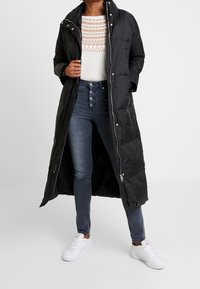 Svart lång pufferjacka med hög krage och tryckknappar, lager-på-lager över en vit tröja med geometriskt mönster och mörka jeans, kombinerat med vita sneakers.