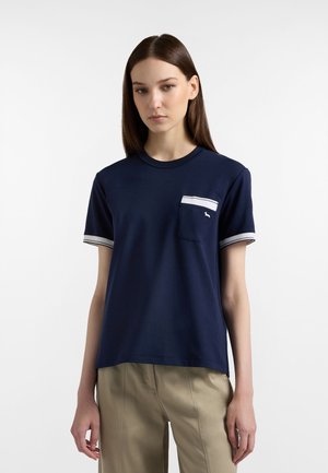 Giovane donna con capelli castani lisci indossa una maglietta blu navy a maniche corte con tasca a righe bianche e pantaloni beige, in piedi davanti a uno sfondo bianco.