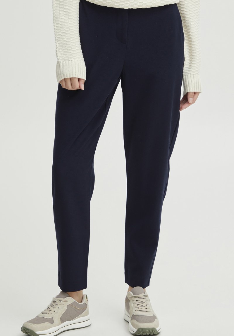 Navy blue pants with a tailored design, featuring a smooth texture and a tapered leg, paired with light-colored sneakers and a cream sweater.