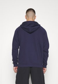 Marineblauwe hoodie met raglanmouwen, een trekkoordcapuchon en een geribde zoom. De stof lijkt zacht en textuur te hebben. Draagt met donkere shorts.