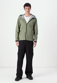 Ελαφρύ παλτό ελιάς με γκρι κουκούλα, μπροστινό φερμουάρ και ρυθμιζόμενους μανικιών, συνδυασμένο με μαύρα cargo παντελόνια και μαύρα υποδήματα.