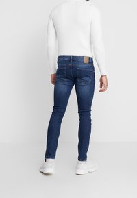 Jeans skinny azul oscuro con bolsillos traseros y una etiqueta de marca marrón, combinados con una camiseta de manga larga blanca y zapatillas blancas.