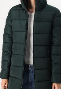 Chaqueta acolchada de longitud media en un tono verde oscuro, llevada abierta sobre una camiseta de rayas blanca y unos vaqueros de tiro alto azules.