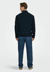 Ein gestrickter navyfarbener Pullover mit hohem Kragen, strukturiertem Muster und gerippten Bündchen, kombiniert mit lässig geschnittenen blauen Jeans.