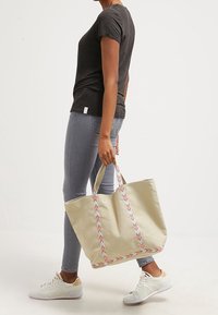 Beige kangaskassi monivärisillä punotuilla olkahihnoilla, yhdistetty harmaisiin kapealahkeisiin farkkuihin ja valkoisiin tennareihin. Yksinkertainen muotoilu ja tilava koko.