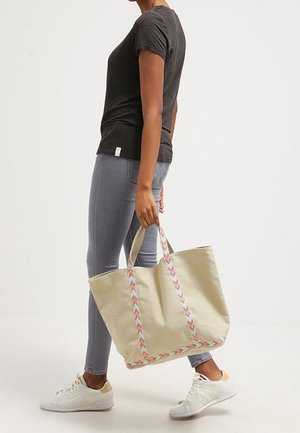 Bolsa de tela beige con asas trenzadas multicolores, combinada con vaqueros pitillo grises y zapatillas blancas. Diseño sencillo y tamaño amplio.